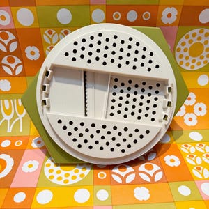 May include: A white and green kitchen gadget with a circular, perforated top and a rectangular grating section. The gadget sits on a green hexagonal base, set against a colorful floral patterned background.