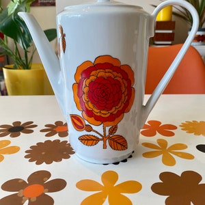 Op de afbeelding: Witte keramische koffiekan met een bloemenmotief. Het ontwerp heeft een grote oranje en rode bloem met gele accenten. De kan heeft een deksel en een handvat.