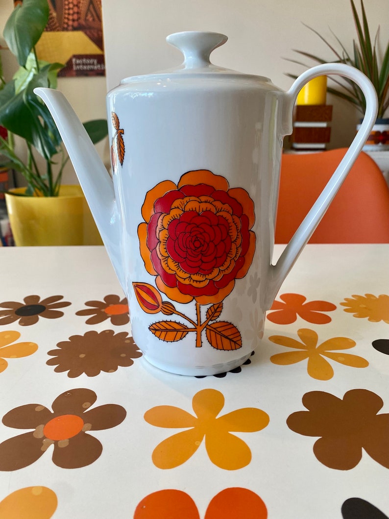 Op de afbeelding: Witte keramische koffiekan met een bloemenmotief. Het ontwerp heeft een grote oranje en rode bloem met gele accenten. De kan heeft een deksel en een handvat.