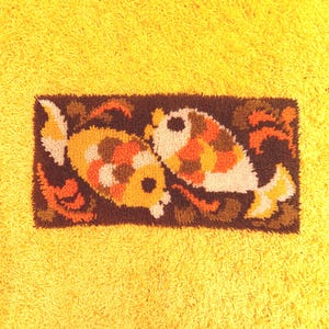 Könnte beinhalten: Ein rechteckiger Teppich im Retro-Stil mit zwei stilisierten Fischen in Gelb-, Orange- und Weißtönen vor einem dunkelbraunen Hintergrund. Der Teppich ist vor einem hellgelben, strukturierten Hintergrund platziert, was einen lebendigen Kontrast erzeugt.