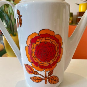 Op de afbeelding: Witte keramische koffiekan met een groot oranje, rood en geel bloemenmotief. De kan heeft een deksel en een tuit.