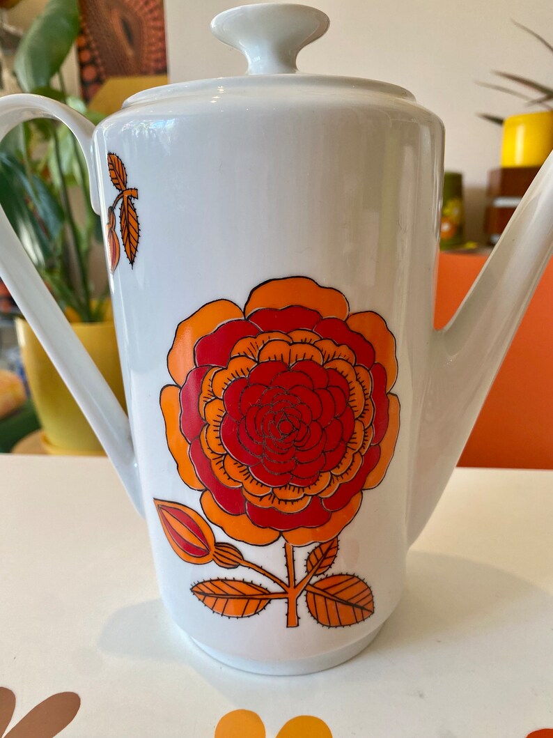 Op de afbeelding: Witte keramische koffiekan met een groot oranje, rood en geel bloemenmotief. De kan heeft een deksel en een tuit.