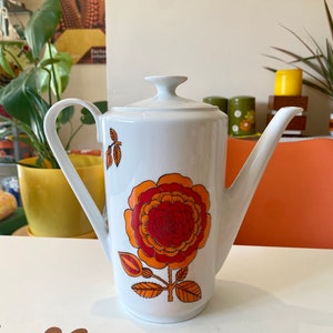 Op de afbeelding: Witte koffiekan van keramiek met een groot bloemenmotief in oranje, rood en geel. De kan heeft een tuit en een handvat.