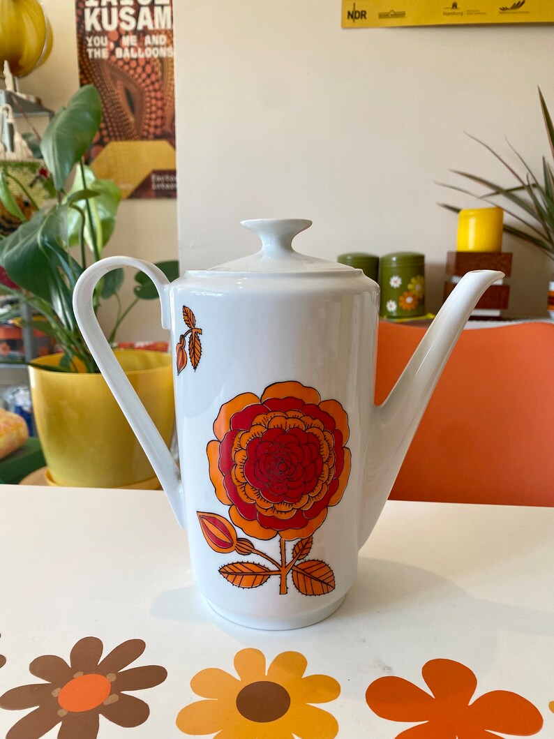 Op de afbeelding: Witte koffiekan van keramiek met een groot bloemenmotief in oranje, rood en geel. De kan heeft een tuit en een handvat.
