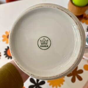 Op de afbeelding: Witte keramische mok met een groene kroon en het woord "KAHLA" gedrukt op de bodem.