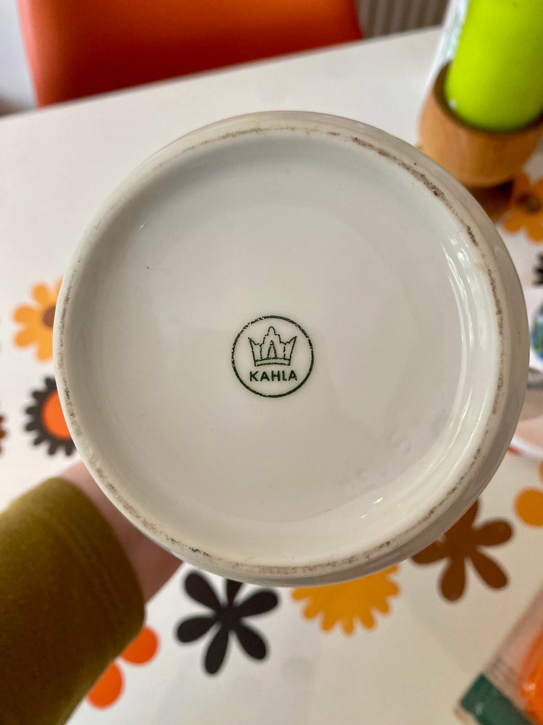 Op de afbeelding: Witte keramische mok met een groene kroon en het woord "KAHLA" gedrukt op de bodem.