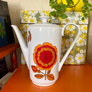 Op de afbeelding: Witte koffiekan van keramiek met een groot bloemenmotief in oranje, rood en geel. De kan heeft een tuit en een handvat.