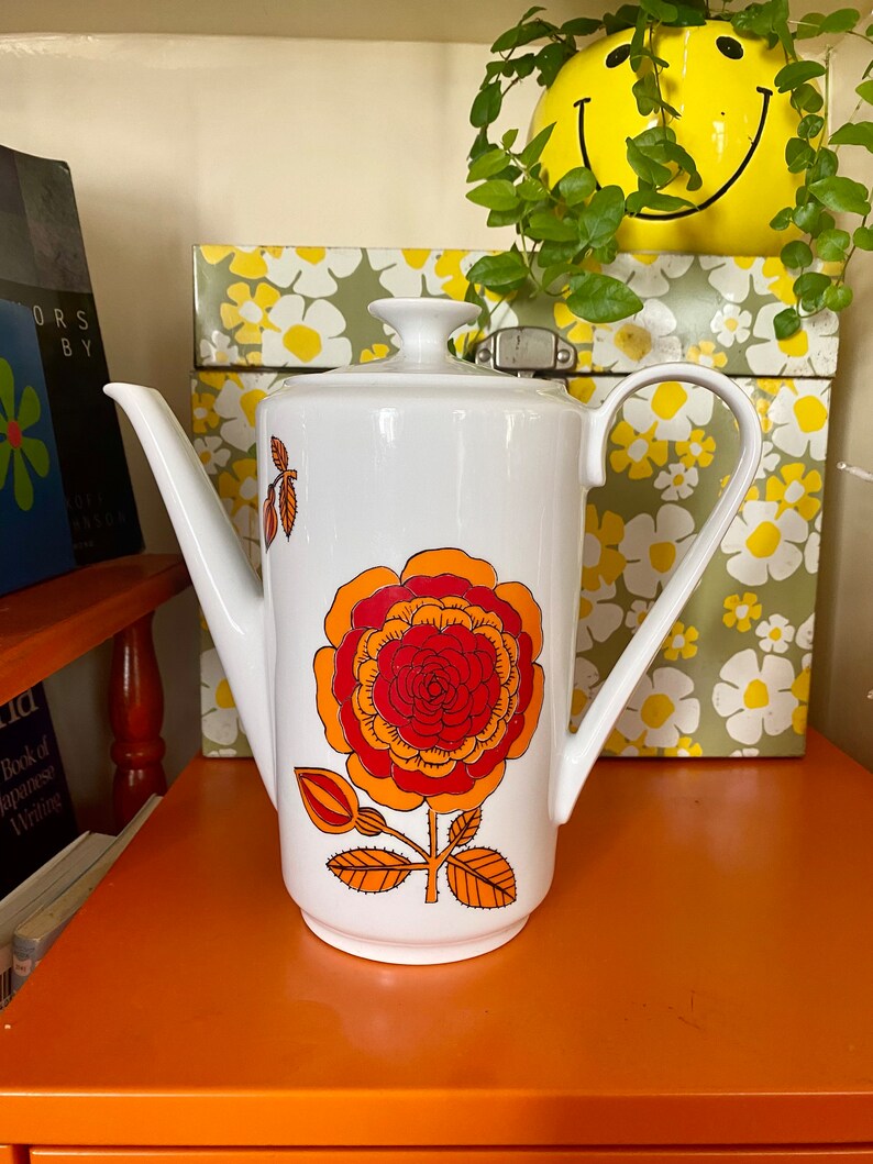 Op de afbeelding: Witte koffiekan van keramiek met een groot bloemenmotief in oranje, rood en geel. De kan heeft een tuit en een handvat.