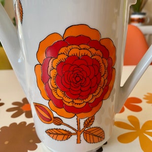 Op de afbeelding: Witte keramische kan met een groot oranje, rood en geel bloemenontwerp. De kan heeft een tuit en een handvat.