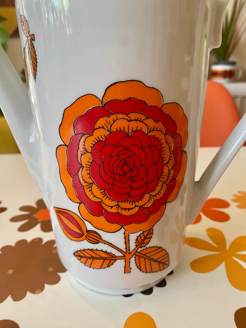 Op de afbeelding: Witte keramische kan met een groot oranje, rood en geel bloemenontwerp. De kan heeft een tuit en een handvat.