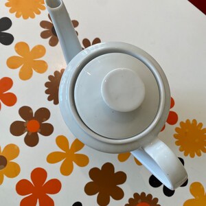 Op de afbeelding: Witte theepot van keramiek met een deksel en een handvat. De theepot staat op een wit oppervlak met een patroon van oranje, bruine en zwarte bloemen.