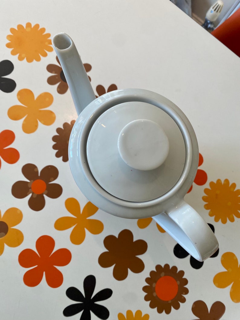 Op de afbeelding: Witte theepot van keramiek met een deksel en een handvat. De theepot staat op een wit oppervlak met een patroon van oranje, bruine en zwarte bloemen.