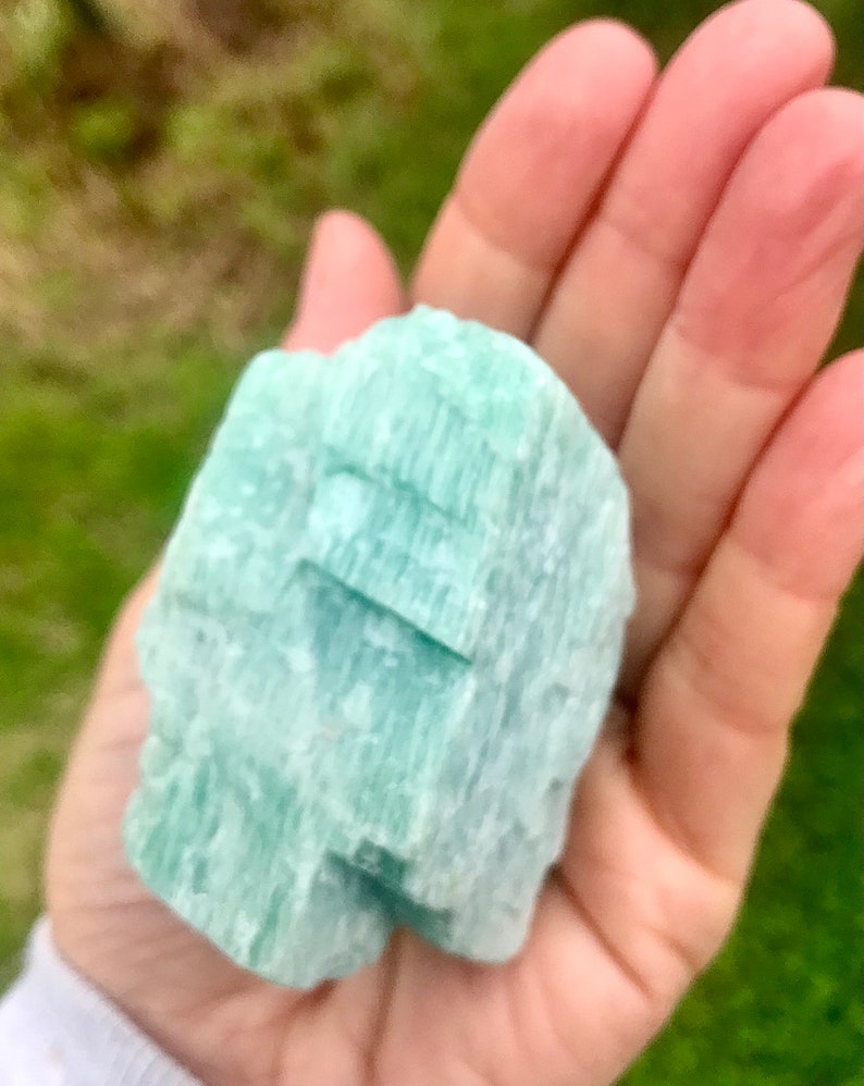 May include: A light green, naturally formed crystal with a striated texture. The crystal is smooth and polished, with a slightly translucent appearance.