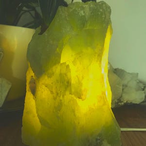 May include: A glowing, rough-hewn, chartreuse-coloured rock lamp. The lamp emits a warm, yellow light from within, creating a soft, ambient glow. The lamp sits on a dark surface, with a power cord visible.