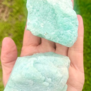 May include: Two large, rough, light green amazonite crystal stones. The stones are in the palm of a hand.
