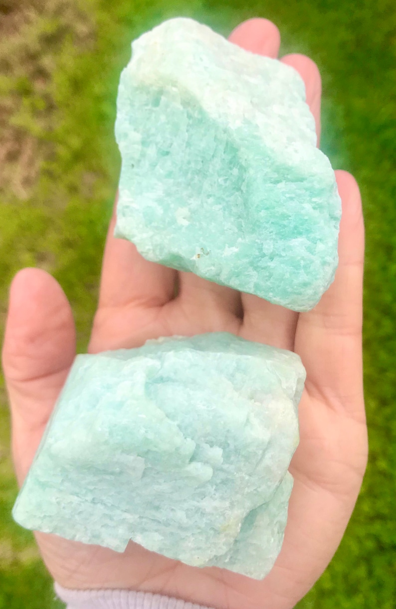 May include: Two large, rough, light green amazonite crystal stones. The stones are in the palm of a hand.