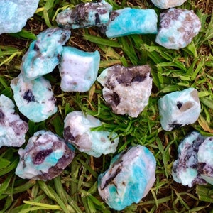 May include: A collection of raw amazonite crystals with a mix of blue, green, white, and black colors. The crystals are rough and have a natural, unpolished look.