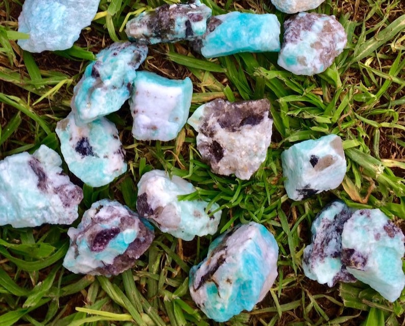 May include: A collection of raw amazonite crystals with a mix of blue, green, white, and black colors. The crystals are rough and have a natural, unpolished look.
