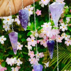 May include: Five amethyst crystal pendulums with silver chains hanging from a hand against a background of pink and white flowers and green leaves.