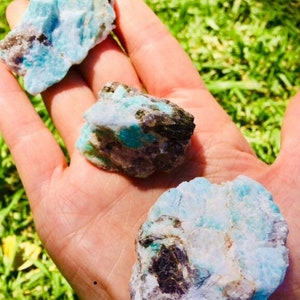 May include: Three rough, natural amazonite stones with a blue-green color and white and black inclusions. The stones are in a hand.