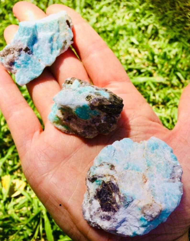 May include: Three rough, natural amazonite stones with a blue-green color and white and black inclusions. The stones are in a hand.