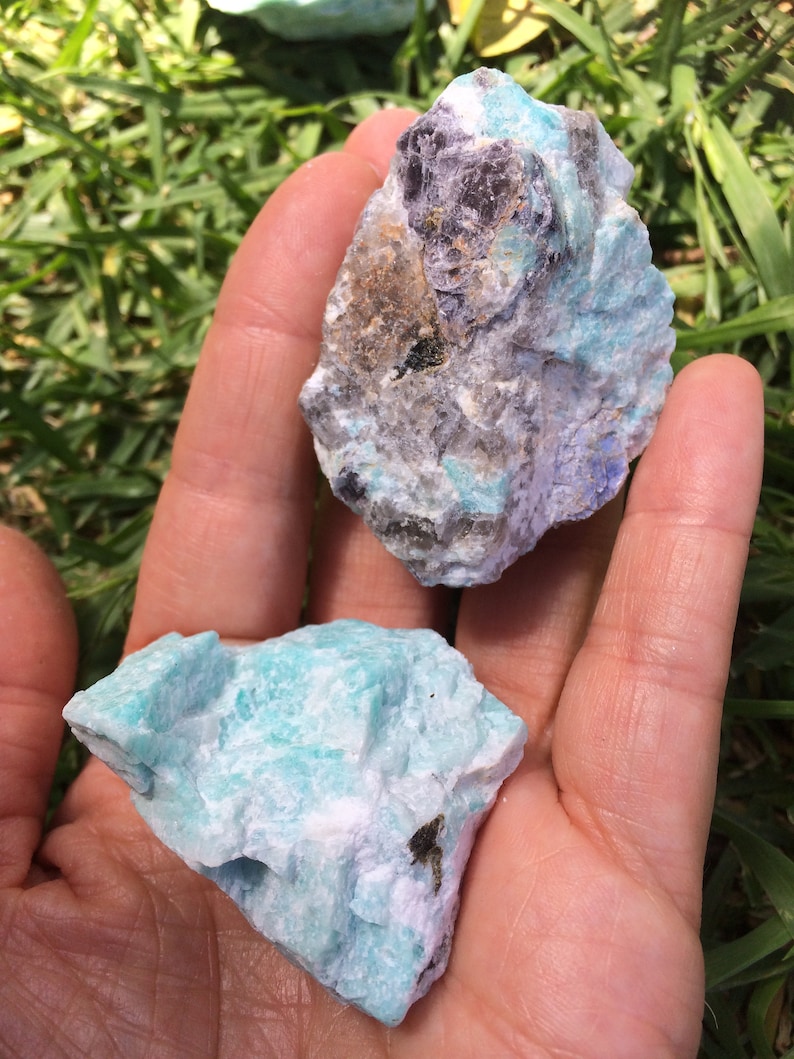 May include: Two rough, natural, green and white amazonite crystals. The larger crystal has a dark gray, almost black, mineral inclusion.