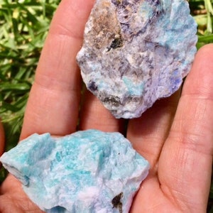 May include: Two rough, natural pieces of Amazonite gemstone. One piece is a light blue-green color with white and gray streaks. The other piece is a darker blue-green color with gray and purple streaks.