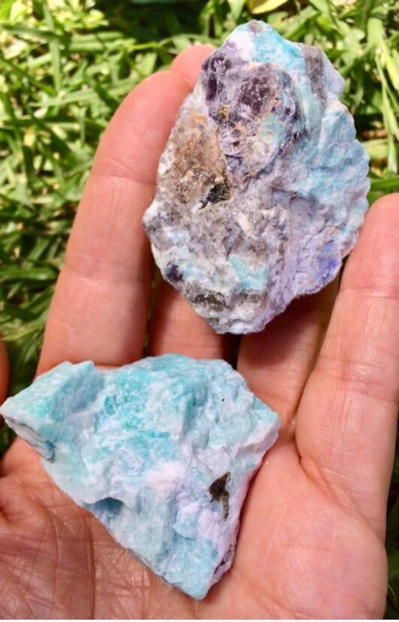 May include: Two rough, natural pieces of Amazonite gemstone. One piece is a light blue-green color with white and gray streaks. The other piece is a darker blue-green color with gray and purple streaks.