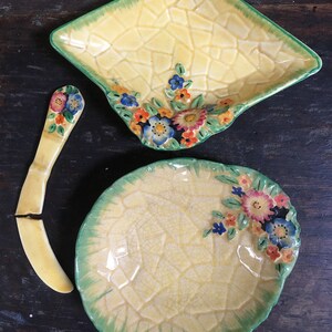Puede incluir: Un juego de tres platos de cerámica vintage con un esmalte amarillo craquelado y un borde verde. Los platos presentan diseños florales con flores rosas, azules y amarillas. Un plato es cuadrado, otro es redondo y el último es un pequeño cuchillo de mantequilla curvo.