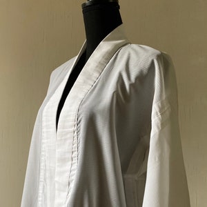 Puede incluir: Un kimono blanco de gasa con un patrón a rayas. El kimono está drapeado sobre un maniquí negro.