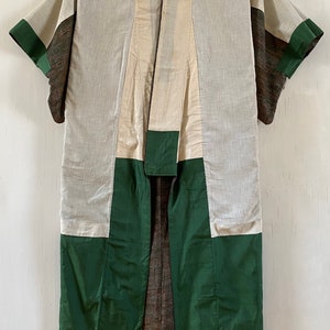 May include: A traditional Japanese robe, or "kimono", with a patchwork design. The kimono is made of a light-colored fabric with green and brown accents. It is hanging on a hanger against a white wall.