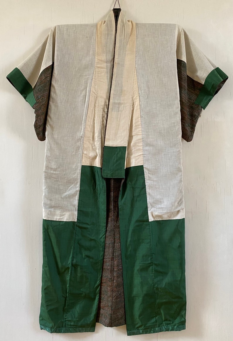 May include: A traditional Japanese robe, or "kimono", with a patchwork design. The kimono is made of a light-colored fabric with green and brown accents. It is hanging on a hanger against a white wall.
