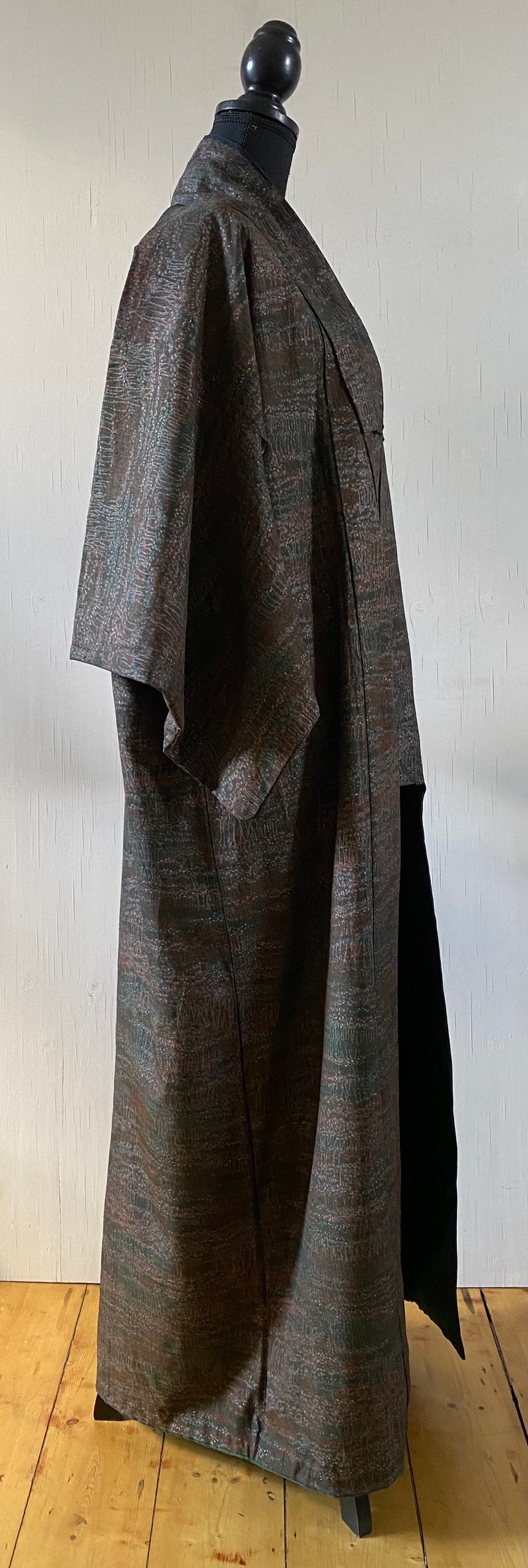 May include: A long, brown and green patterned coat with a short sleeve. The coat is on a mannequin and is standing on a wooden floor.
