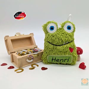 Könnte beinhalten: Grünes, flauschiges Monster-Plüschtier mit gestickten Augen, einem lächelnden Mund und einem roten Herzen. Das Spielzeug hat ein Namensschild mit der Aufschrift "Henri".