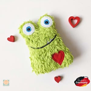 Könnte beinhalten: Grünes, flauschiges Monster-Plüschtier mit großen blauen Augen und einem roten Herzen auf der Brust. Das Spielzeug ist handgefertigt in Deutschland.
