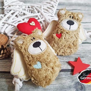 Könnte beinhalten: Zwei braune Teddybär-Anhänger mit weißen Flügeln und roten Herzen. Die Bären sind handgefertigt und haben den Text "Handmade in Germany" auf einem roten Stern.