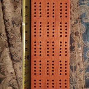 May include: A rectangular wooden cribbage board with a natural light brown finish and multiple rows of drilled peg holes. A yellow Stanley measuring tape is positioned along the left side, indicating the board is approximately 53 centimetres long.