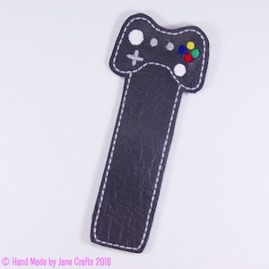 May include: A grey leather bookmark shaped like a video game controller with white and coloured buttons. The bookmark has white stitching around the edges.