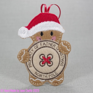 May include: A felt gingerbread man ornament with a red Santa hat, brown eyes, and pink cheeks. The gingerbread man is holding a round button with the text "PROPERTY OF FATHER CHRISTMAS NORTH POLE".