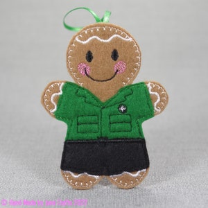May include: A gingerbread person ornament wearing a green uniform with a scout emblem. The ornament has a brown face with pink cheeks and a white smile.