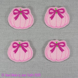 May include: Four identical light pink pumpkin-shaped felt appliqués, each featuring a bright magenta embroidered bow at the top and vertical stitched lines. The text "© Hand Made by Jane Crafts 2026" is printed in magenta at the bottom left.