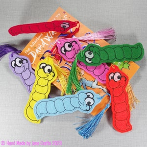May include: A set of colourful felt worm bookmarks with embroidered eyes and a tassel. The bookmarks are arranged on a book with the text "the hilarious David Walliams" visible.