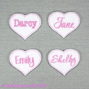 May include: Four white heart-shaped fabric patches with pink embroidered names. The names Darcy, Jane, Emily, and Shelby are stitched in a cursive font. Each patch features a pink stitched border around the edge of the white heart shape.