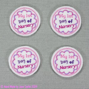 May include: Four white, round, felt badges with pink and purple embroidery. Each badge says "My 1st Day of Nursery" in a pink and purple font.