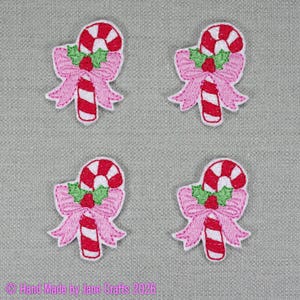 May include: Four embroidered patches, each featuring a red and white striped candy cane adorned with a light pink bow and green holly leaves with red berries. The patches are arranged in a grid pattern on a textured grey fabric background.