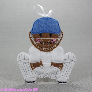 May include: A felt ornament of a cricket player wearing a blue helmet, white uniform, and white pads. The player is in a catching position with a brown face and pink cheeks.