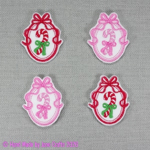 May include: Four embroidered patches featuring oval frames with bows at the top and candy cane designs in the centre. Two patches have red frames with red and green candy canes, and two have pink frames with pink candy canes on a grey fabric background.