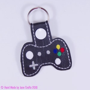 May include: A black leather video game controller keyring with white stitching and colourful buttons. The keyring has a silver key ring.