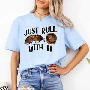 May include: A light blue t-shirt featuring a graphic of a brown pill bug and a rolled-up pill bug with the black text "JUST ROLL WITH IT" printed in the center. The design shows the insect in both its extended and curled-up defensive positions.