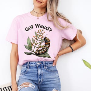 May include: A light pink t-shirt with the text "Got Weed?" in black. The shirt features a colorful illustration of a caterpillar on a plant with green leaves and orange berries. The shirt is made of a soft material.